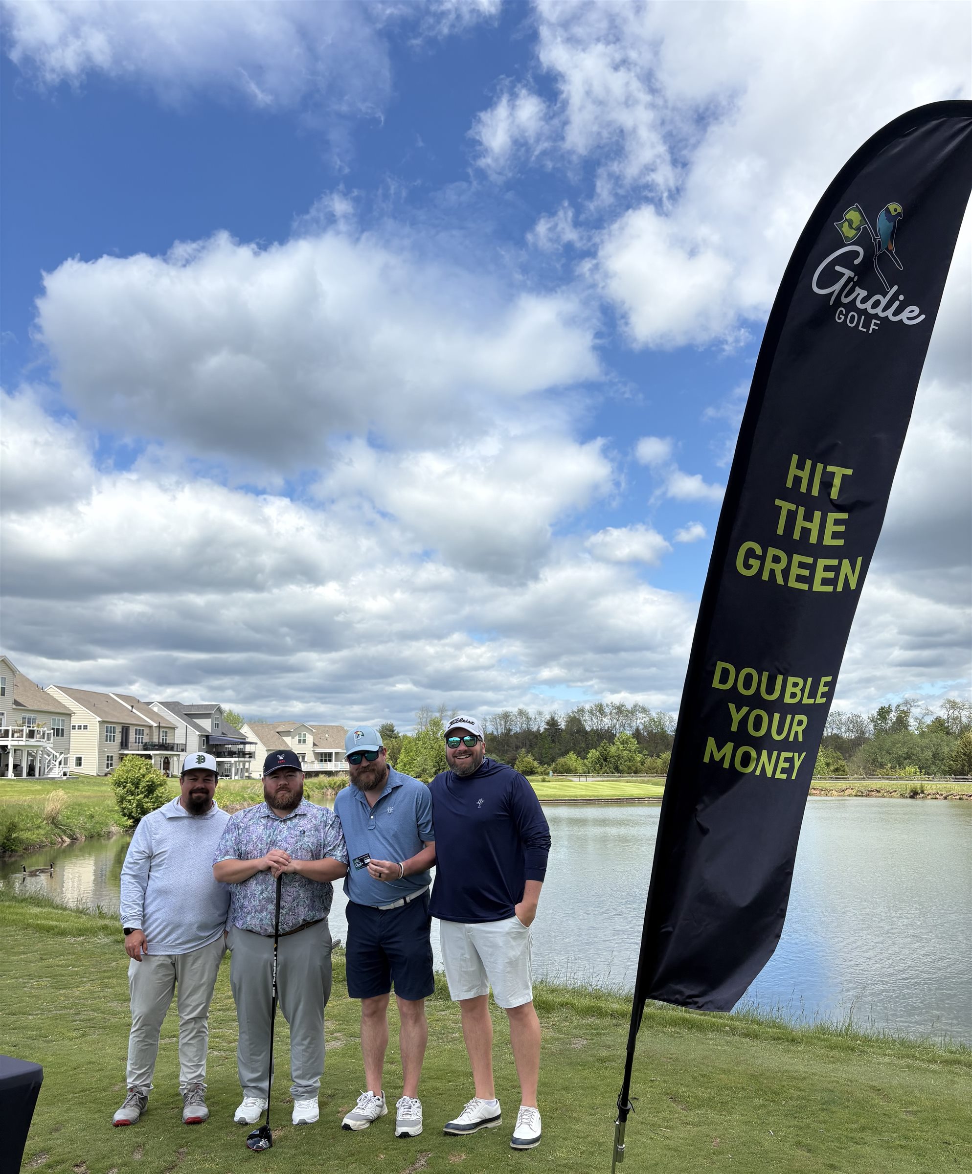 Four golfers beside the Girdie Golf challenge banner on a par-3 tee overlooking water
