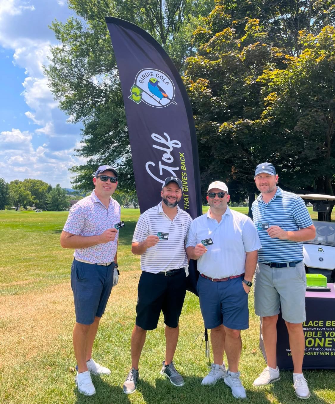 Four golfers holding up pro shop gift cards they won, beside the Girdie Golf challenge banner on a par-3 tee